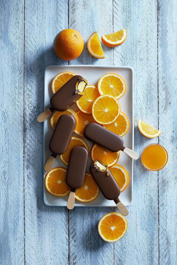 Schokoladen-Orangen-Popsicle-Eis auf einem weißen Teller mit frisch geschnittenen Orangenscheiben auf blauem Holzhintergrund