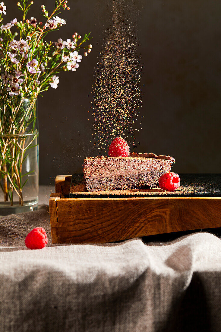 Bestäubtes Kakaopulver fällt auf ein Stück Lissabonner Schokoladenkuchen mit Himbeeren als Garnitur auf einem Tisch mit Blumen im Hintergrund.