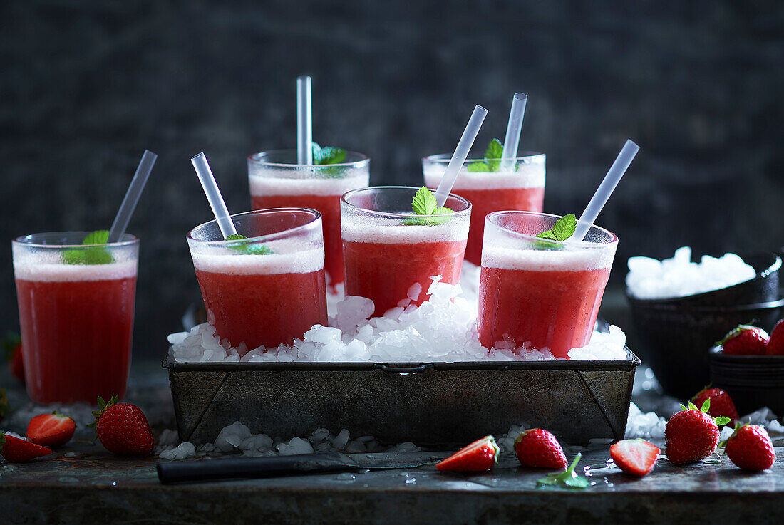 Homemade iced strawberry drinks on crushed ice in a metal container decorated with fresh strawberries on a gray metal background