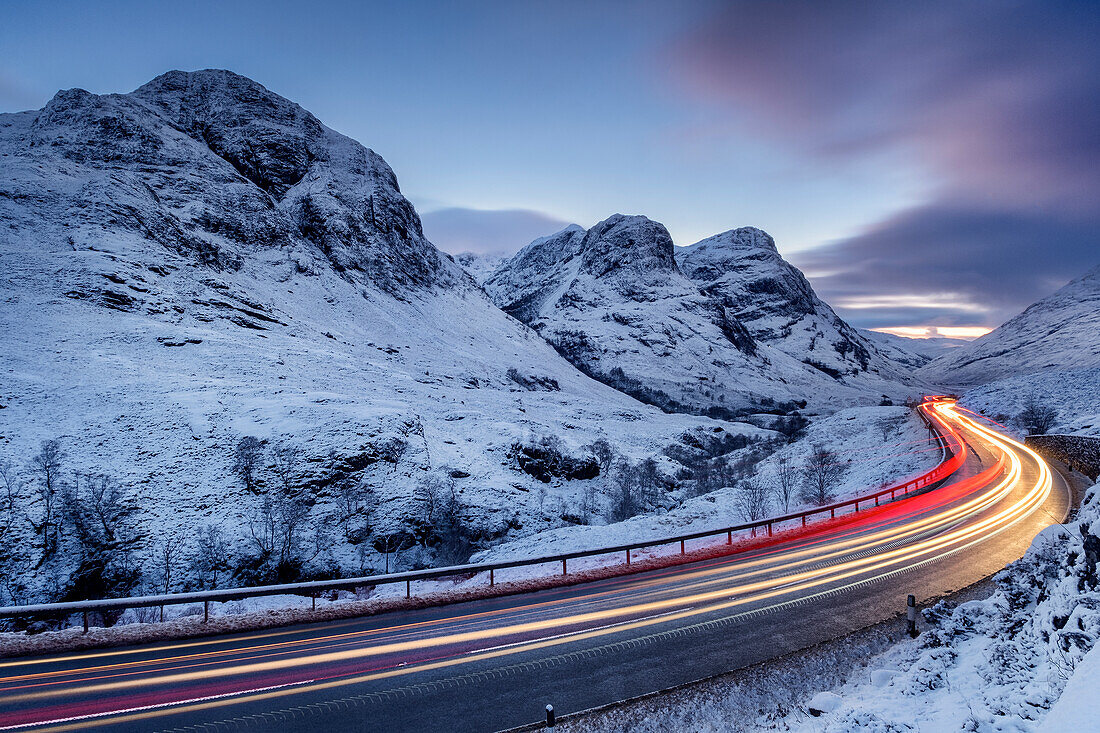 Verkehr auf der A82, vorbei an den schneebedeckten Three Sisters of Glencoe, Aonach Dubh, Beinn Fhada und Gearr Aonach, bei Nacht im Winter, Glencoe, Schottische Highlands, Schottland, Vereinigtes Königreich, Europa