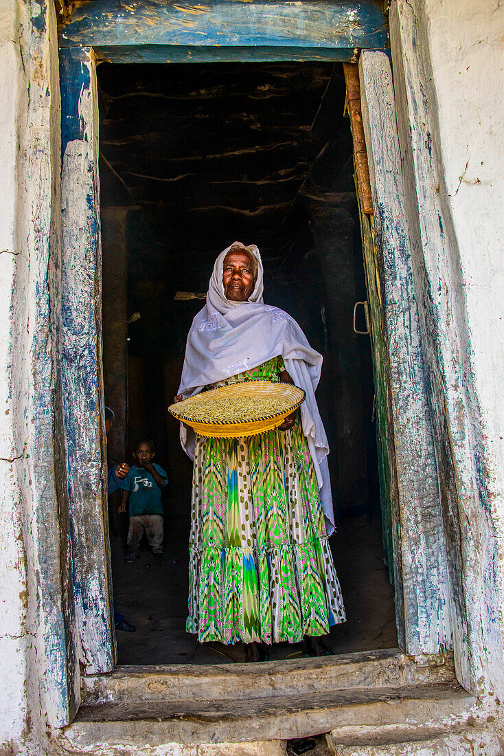 Freundliche alte Frau steht mit einem Korb voller Mais in einem Türrahmen, bei Keren, Eritrea, Afrika