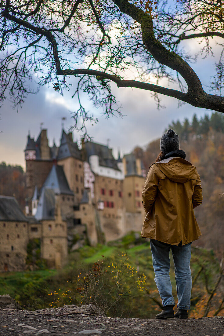 Back view of young woman with yellow jacket looking at Eltz medieval historic castle in an autumn landscape with trees at sunrise, Wierschem, Rhineland-Palatinate, Germany, Europe