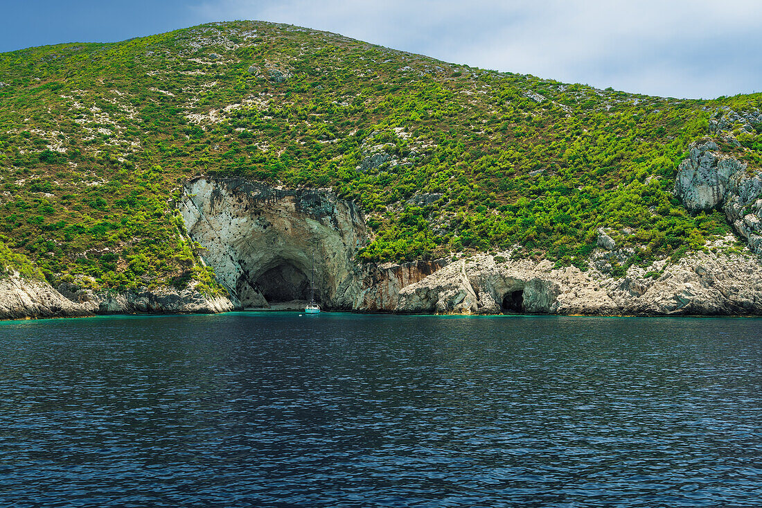 Vertäute Yacht vor einer Höhlenformation auf einem felsigen Hügel mit grüner Plantage, die sich bis zum Meer erstreckt, Zakynthos, Ionische Inseln, Griechische Inseln, Griechenland, Europa