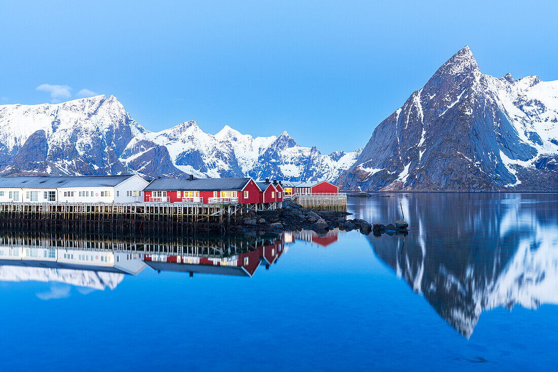 Abenddämmerung über dem Fischerdorf und dem ruhigen Wasser des Fjordes, umgeben von schneebedeckten Bergen, Hamnoy, Reine, Moskenes, Lofoten, Norwegen, Skandinavien, Europa