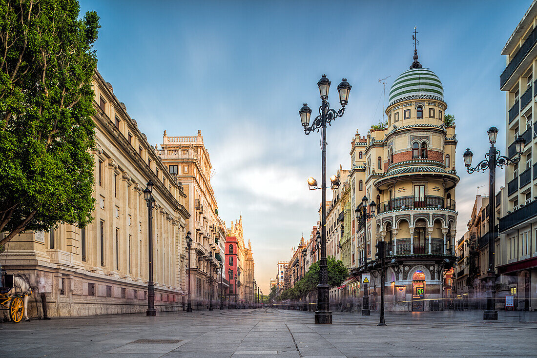 A long exposure shot captures the empty Avenida de la Constitucion in Seville, Spain at sunset. The image shows the street lined with buildings and streetlights.