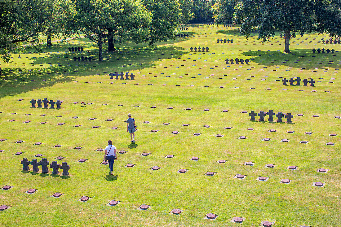 Deutscher Soldatenfriedhof in der Normandie, Frankreich, mit Besuchern, die unter sonnigem Himmel über die Gräberreihen nachdenken.