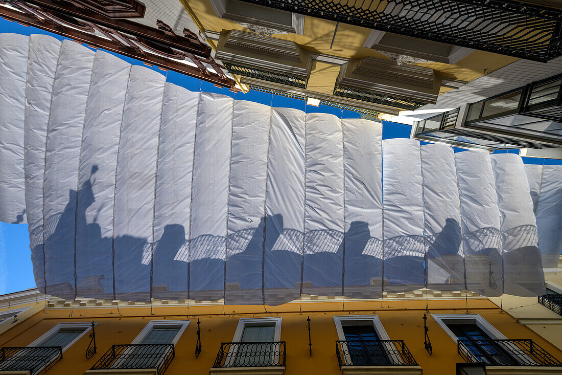 A white shade awning installed above a street in the center of Sevilla to protect against the summer heat. Colorful building facades are visible.