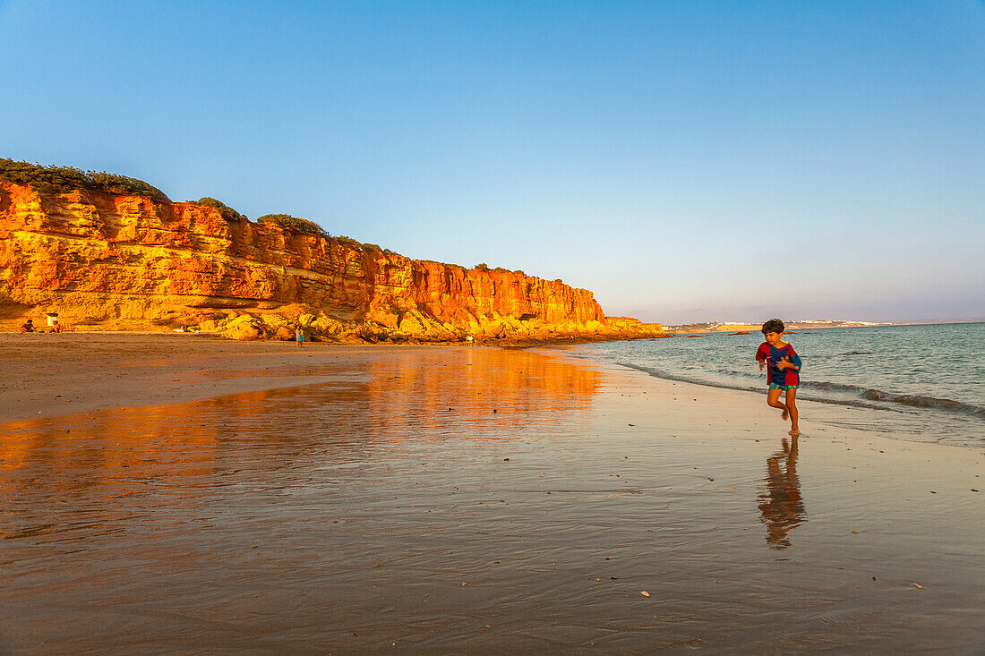 A child running on the sandy beach of Cala en Cabo Roche, Cadiz, Andalusia, Spain, during sunset. Majestic cliffs and calm waters surround the scene.