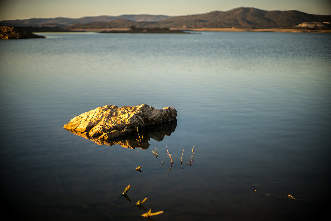 Ruhige Abendaufnahme des Stausees Puente Nuevo mit einer ruhigen Wasseroberfläche, einem Felsen und fernen Bergen in Villaviciosa de Cordoba, Andalusien, Spanien.