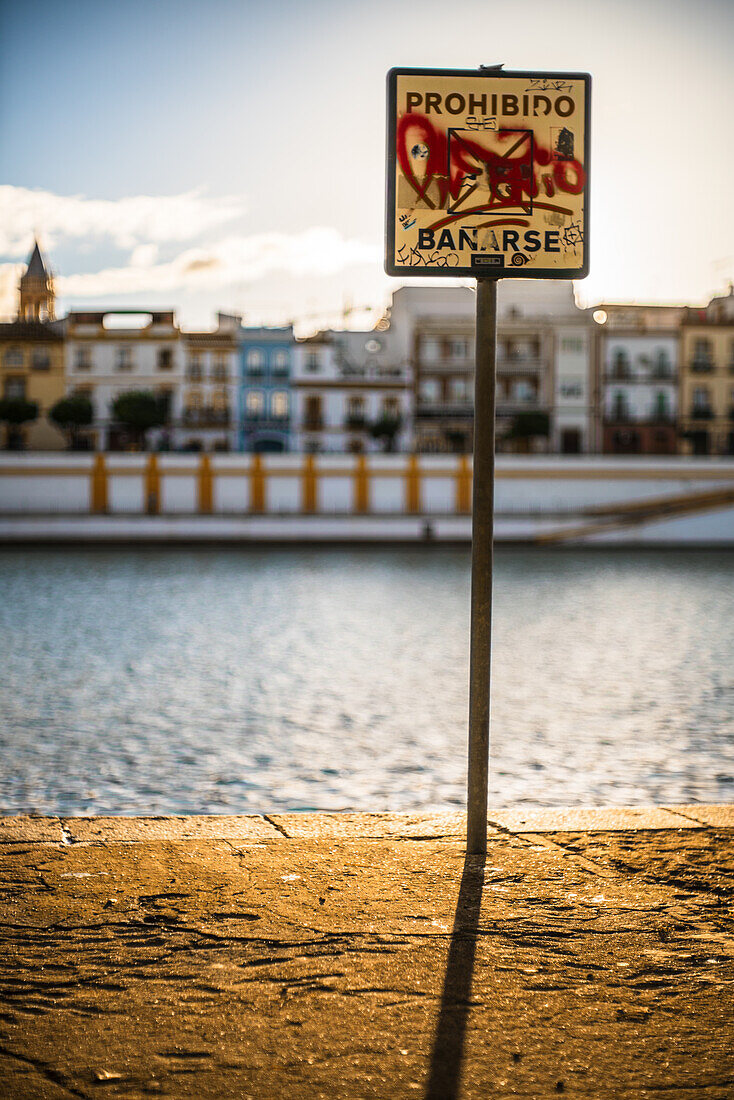 A graffiti-covered 'Prohibido Bañarse' sign on the riverbank in Sevilla, España, with colorful buildings in the background.