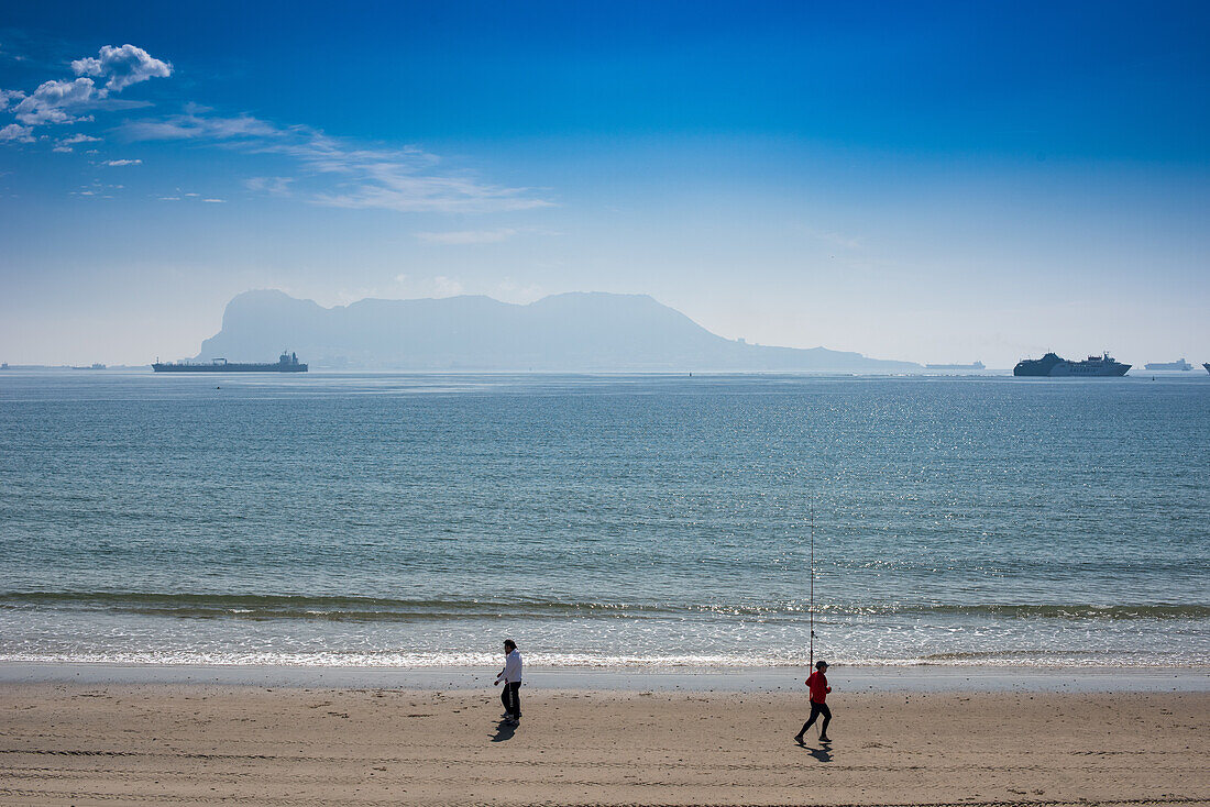 People walking and running along El Rinconcillo beach in Algeciras, Spain, with cargo ships and Gibraltar in the distant background.