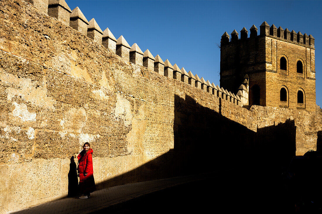 Frau in rotem Mantel vor der historischen Muralla de la Macarena in Sevilla. Sonnenbeschienene alte Mauer mit Schatten.