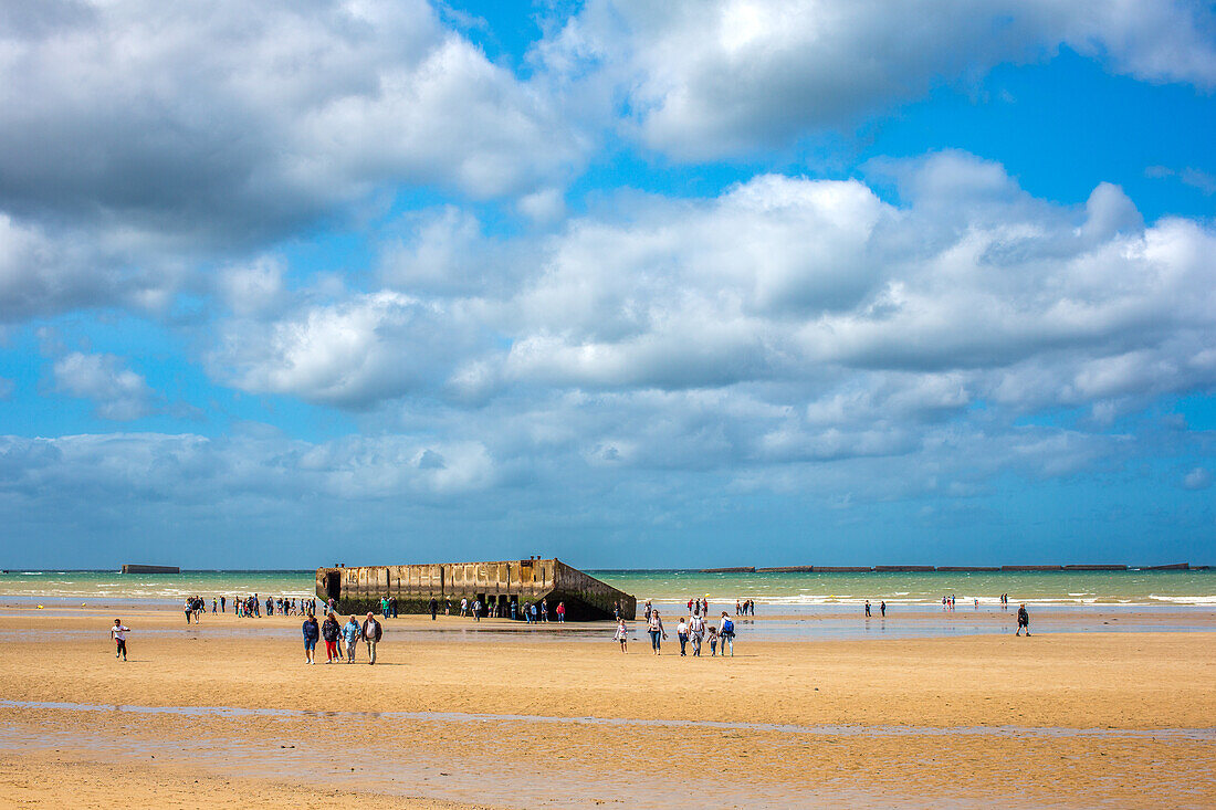 Touristen erkunden den Goldstrand in Arromanches, Normandie, Frankreich, mit den Überresten des Hafens von Mulberry B im Hintergrund.