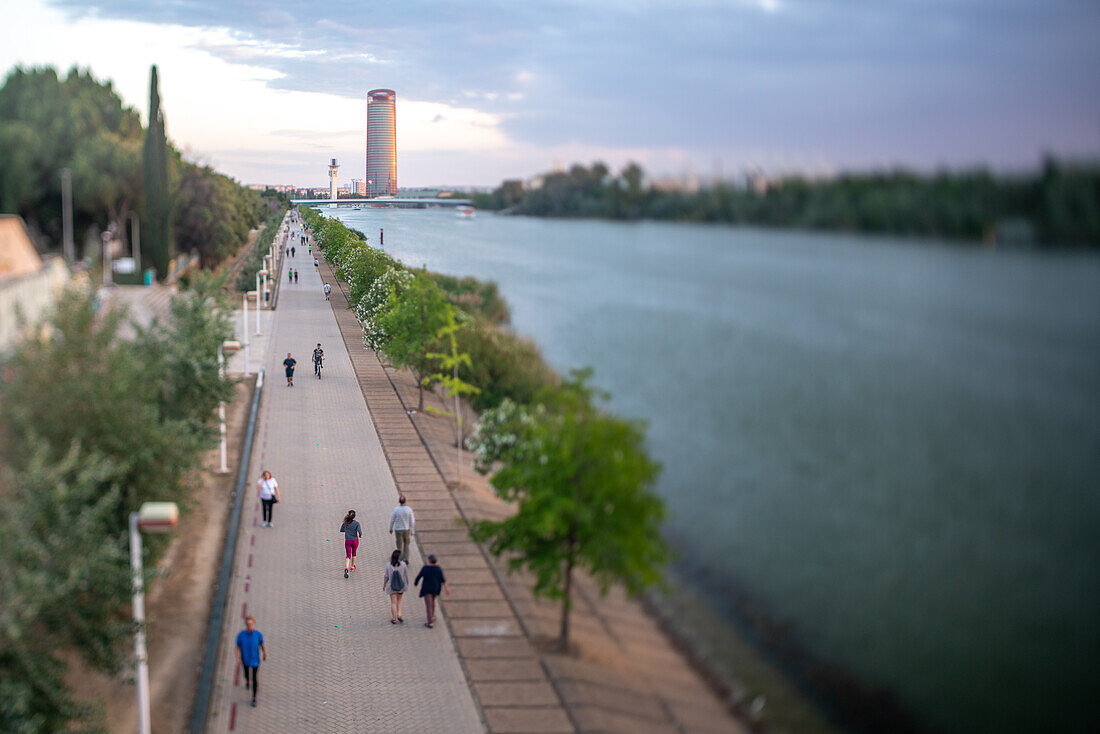 People strolling and cycling along a riverside walkway with Torre Sevilla in the background during dusk. A serene urban scene in Sevilla.