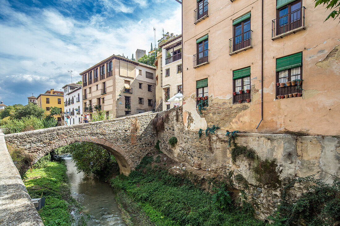 Blick auf historische Gebäude und eine Steinbrücke über den Fluss Darro in Granada, Spanien, die den Charme der alten Welt und die malerische Umgebung widerspiegeln.