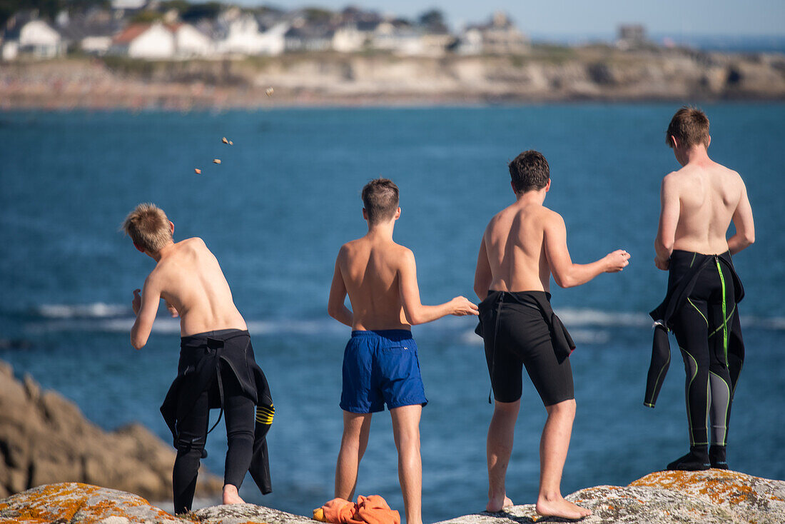 Eine Gruppe von Jungen genießt einen sonnigen Tag, indem sie von einer Klippe in Ouistreham, Bretagne, Frankreich, Steine ins Meer werfen.