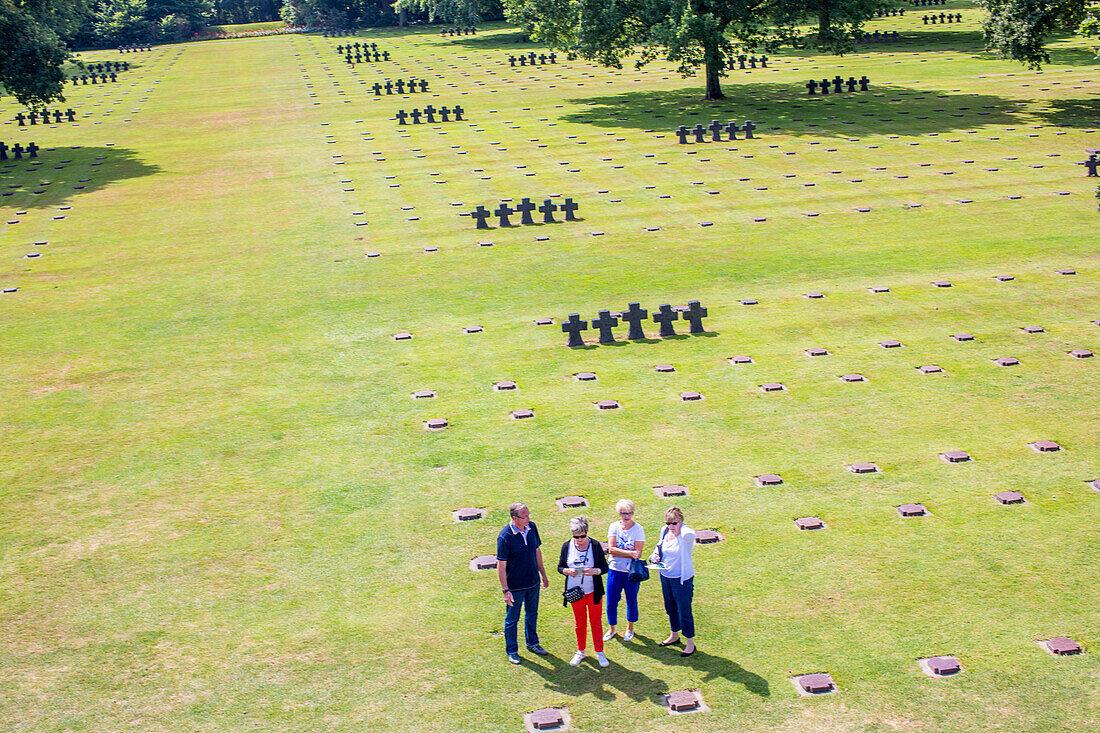 A group of visitors stands at the German military cemetery in Normandy, France, paying respect amidst rows of graves.