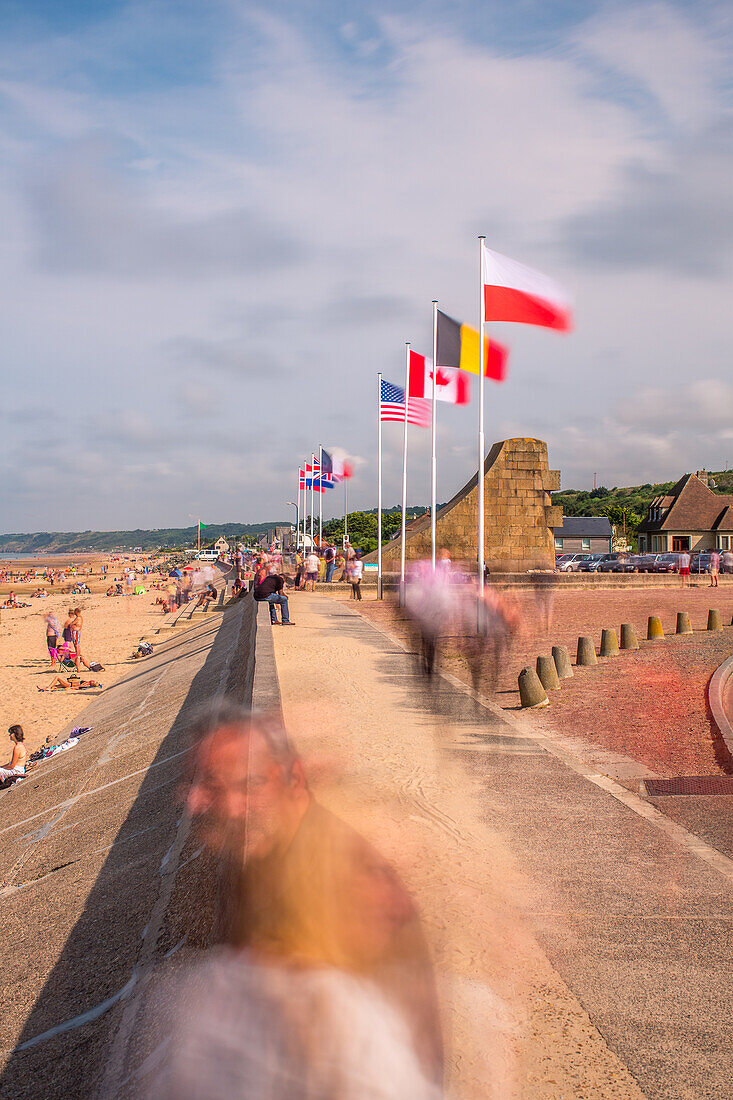 Long exposure photograph of the promenade at Omaha Beach in Normandy, France, featuring various national flags and beach visitors.