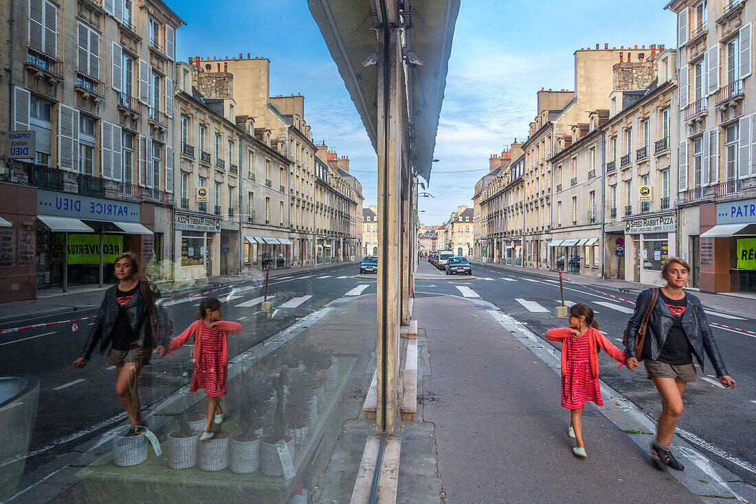 A mother and daughter walking hand in hand along a quiet street in Caen, Normandy, France. The reflection in the window captures their bond and the charming architecture around.