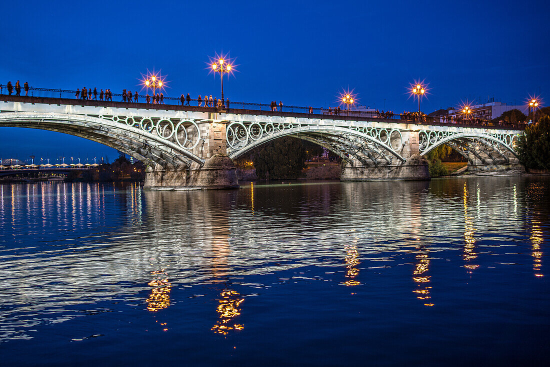 Nighttime view of the illuminated Triana Bridge over the Guadalquivir River in Seville, Spain. Reflections on the water and city lights create a serene atmosphere.