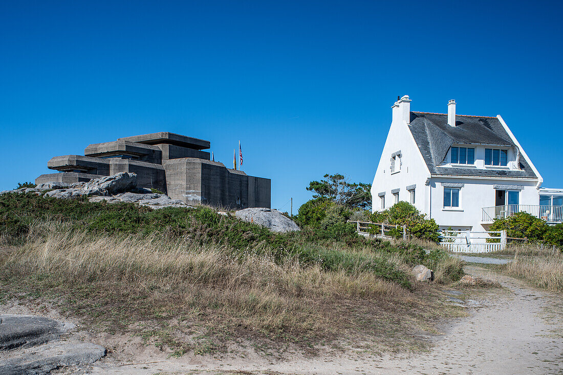 Der große Bunker der ehemaligen deutschen Kommando- und Feuerleitstelle in Ouistreham, Bretagne, Frankreich, neben einem nahe gelegenen Haus unter einem klaren blauen Himmel.