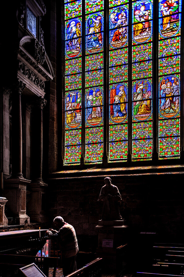 A man prays by the stained glass windows in Collegiale Saint Aubin, Guerande, France. The colorful windows depict religious scenes.