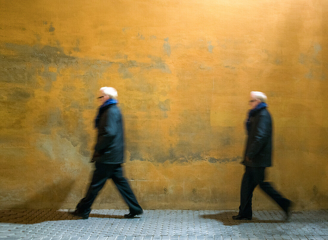 Man Pursuing Himself past a vibrant yellow wall in Sevilla, España. Captures the motion and energy of city life.