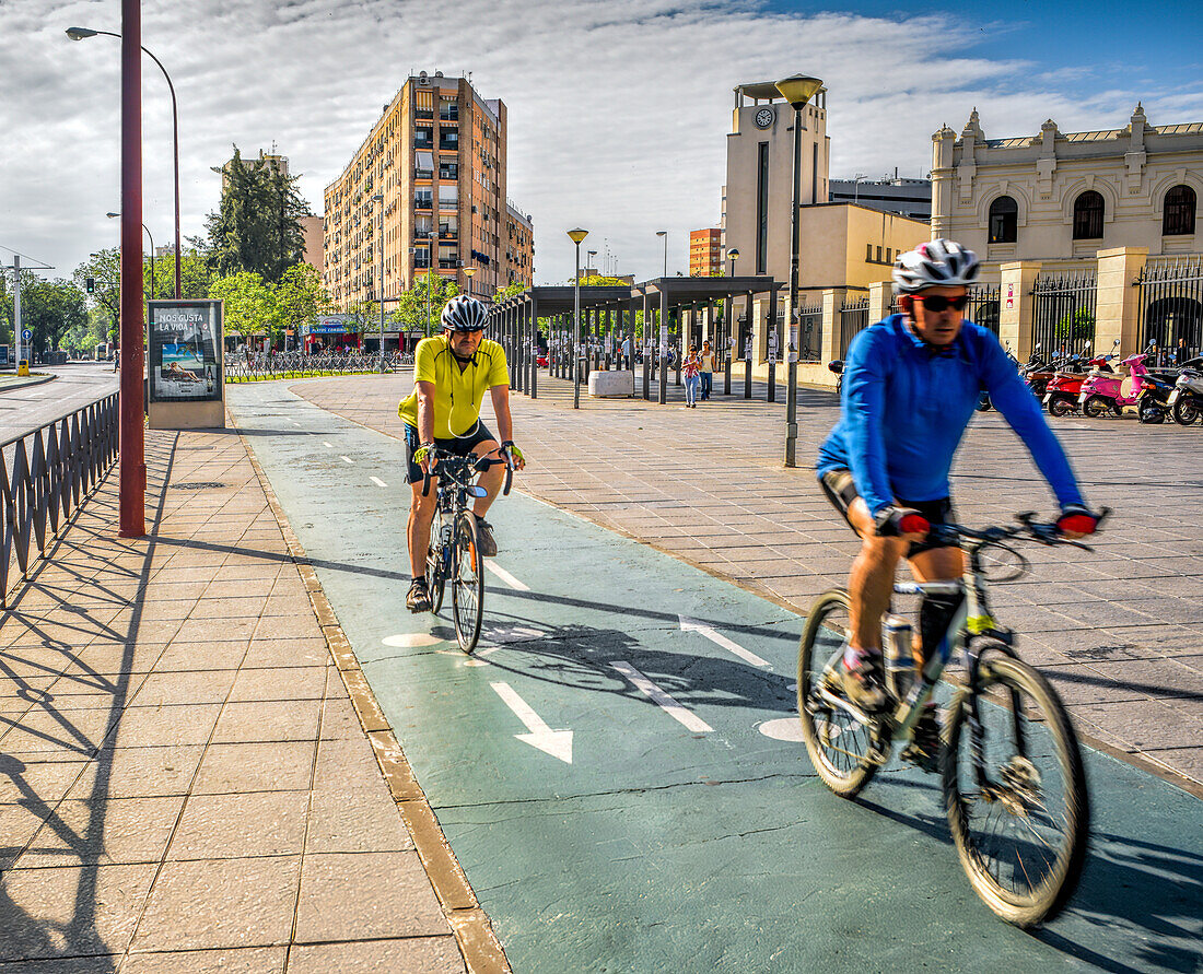 Two cyclists ride through the Ramon y Cajal area in Seville, Andalusia, Spain on a sunny day, enjoying the city's urban landscape.