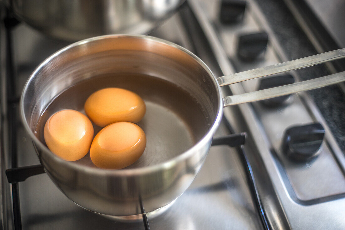 Nahaufnahme eines Topfes auf einer Herdplatte, in dem drei Eier in Wasser kochen.