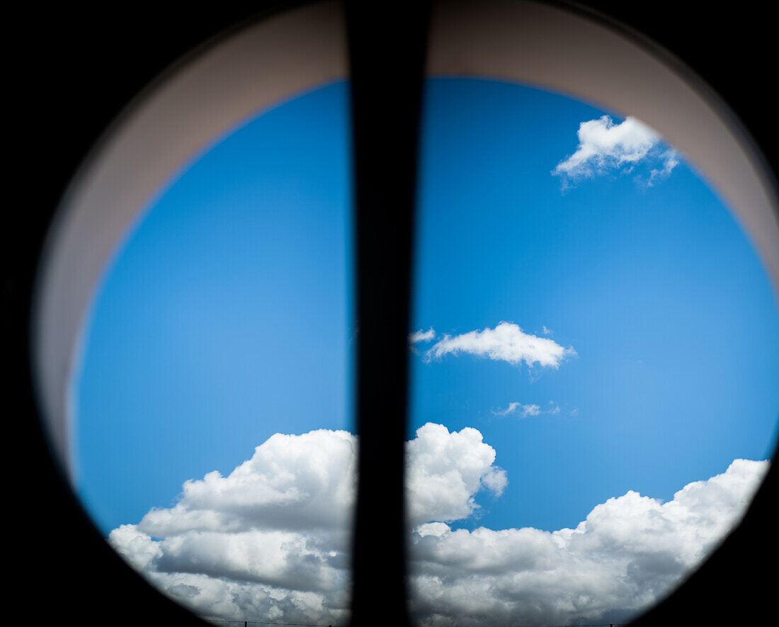 Ein strahlend blauer Himmel mit flauschigen weißen Wolken, umrahmt von einem runden Fenster, das eine heitere und friedliche Stimmung einfängt.