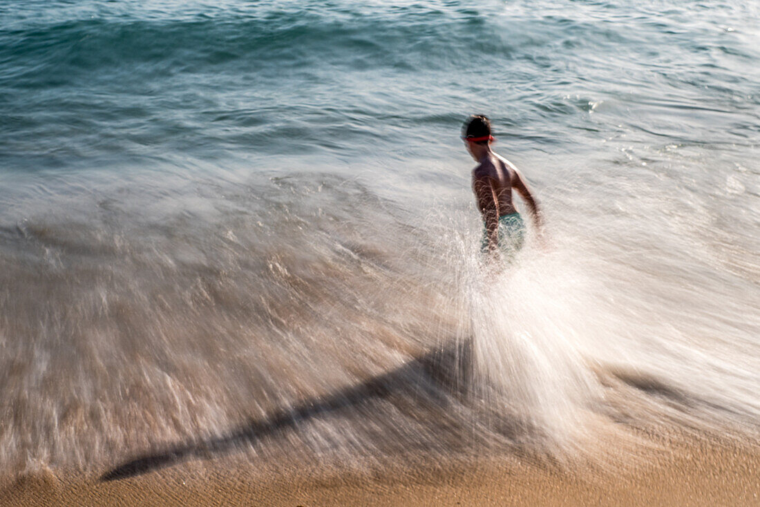 Ein lang belichtetes Foto eines Jungen, der am Strand von Zahora, Cadiz, Spanien, in den Wellen spielt. Die Bewegungsunschärfe vermittelt ein Gefühl von Energie und Bewegung.