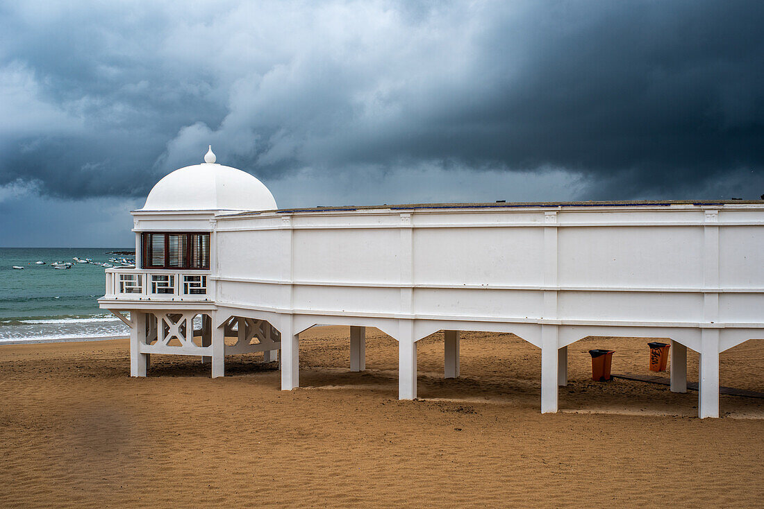 Das kultige Badehaus Balneario De La Palma am Strand Playa De La Caleta in Cádiz, Andalusien, Spanien, unter einem dramatisch bewölkten Himmel.