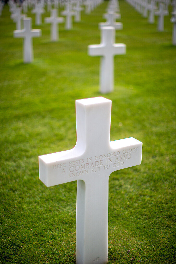 White cross for an unkown soldier at the American military cemetery in Normandy, France, honoring fallen soldiers with rows of graves on green grass.