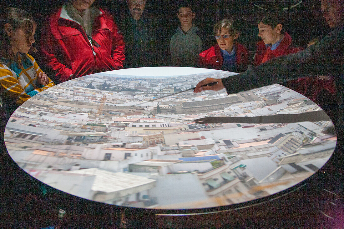 Visitors examine an image of downtown Sevilla projected inside a camera obscura exhibit, appreciating the detailed cityscape.