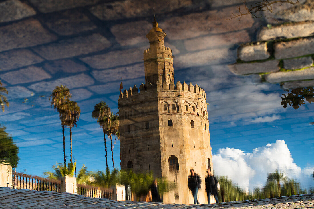 Stunning view of the Torre del Oro reflecting in a puddle after rain in Seville, Spain. Clear blue sky enhances the vibrant colors and historical detail.