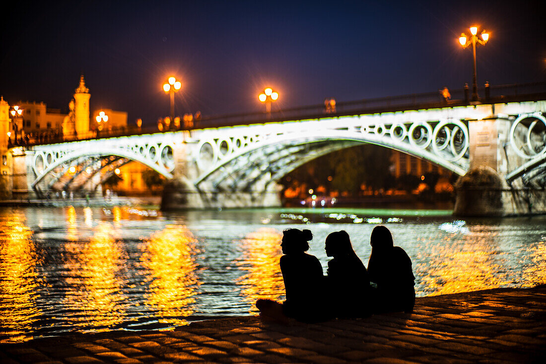 A group of friends enjoying an evening by the riverside near a beautifully illuminated Triana bridge in Seville, Spain. Captured with a Leica Noctilux lens.