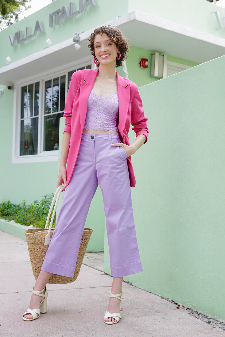 Young woman in lavender-coloured combination with pink blazer in front of mint-green building