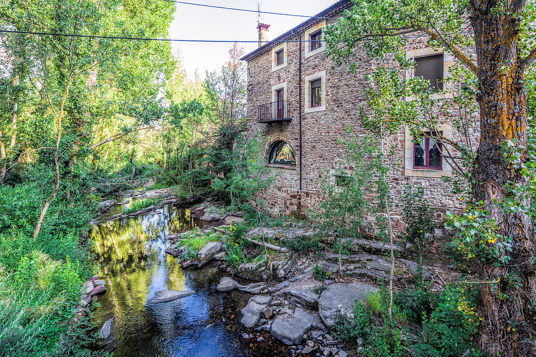 The serene river Tera flows gently past a rustic stone building surrounded by lush greenery in Almarza, Soria, Spain.