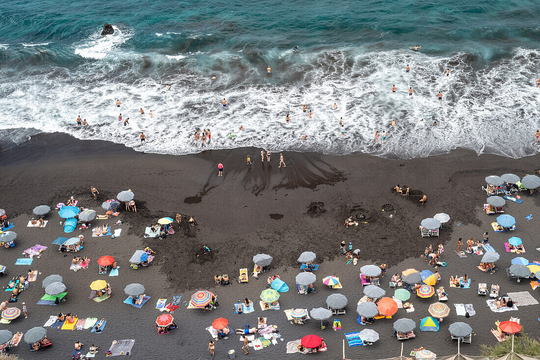 Aerial view of Playa del Bollullo in Tenerife with beachgoers, black sand, and crashing ocean waves. Located in La Orotava, Canary Islands, Spain.