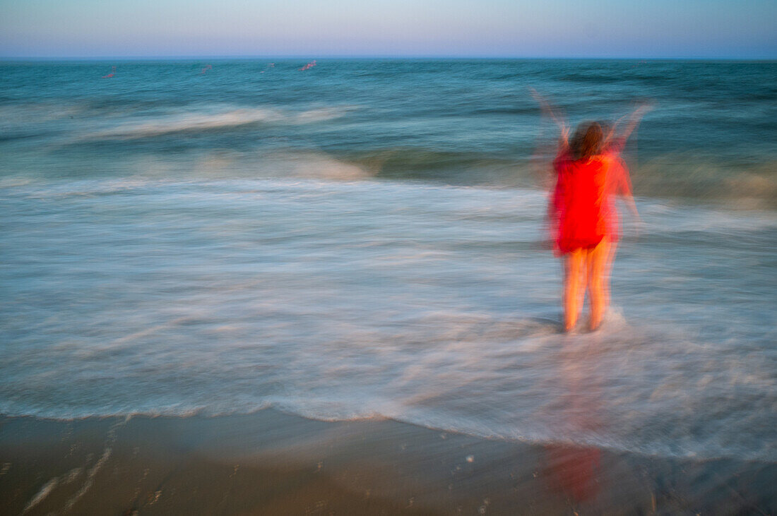 Eine lebhafte Langzeitbelichtung eines Mädchens in einem roten Kleid, das am Strand spielt und die Bewegung der Meereswellen einfängt