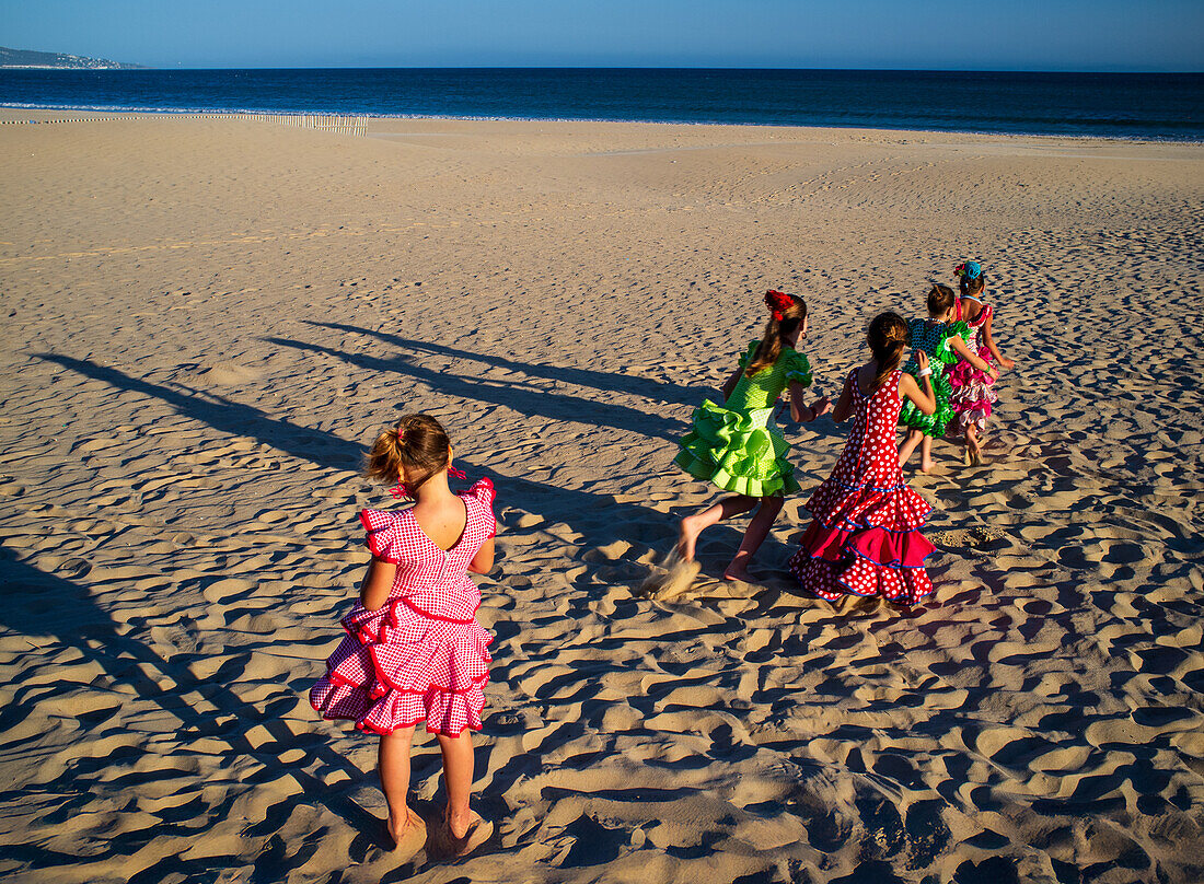 Vibrant scene of young girls in colorful flamenco dresses enjoying the beach in Barbate, Cadiz during local festivities, showcasing cultural traditions.