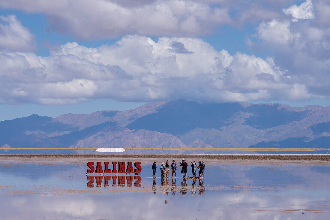 Tourists by the metal sign on the Salinas Grandes salt flats on the altiplano in Argentina. Recent rains left a shallow sheet of water on the flats.