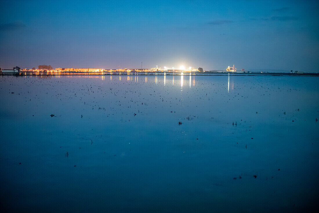 Scenic view of flooded fields under winter skies in Isla Mayor, Sevilla, Spain, prepared for rice cultivation. Evening lights reflect on the water surface, creating a tranquil atmosphere.