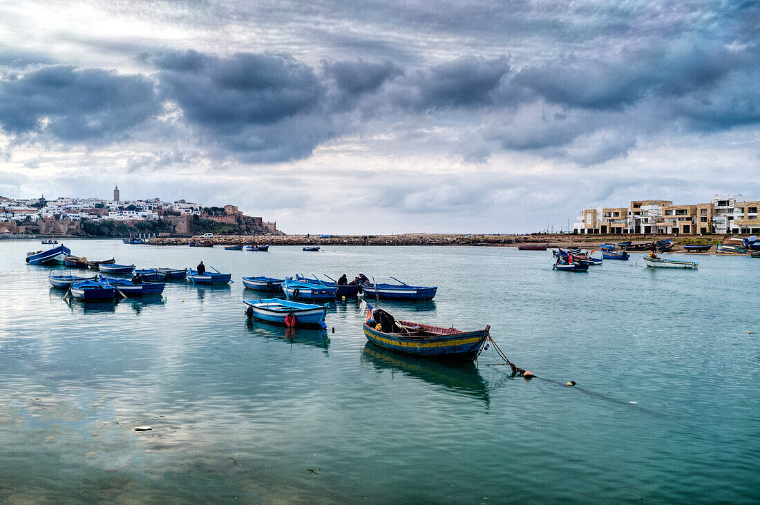 Colorful fishing boats float on the calm waters of Bou Regreg River as dusk settles over Rabat, Morocco, creating a serene atmosphere.