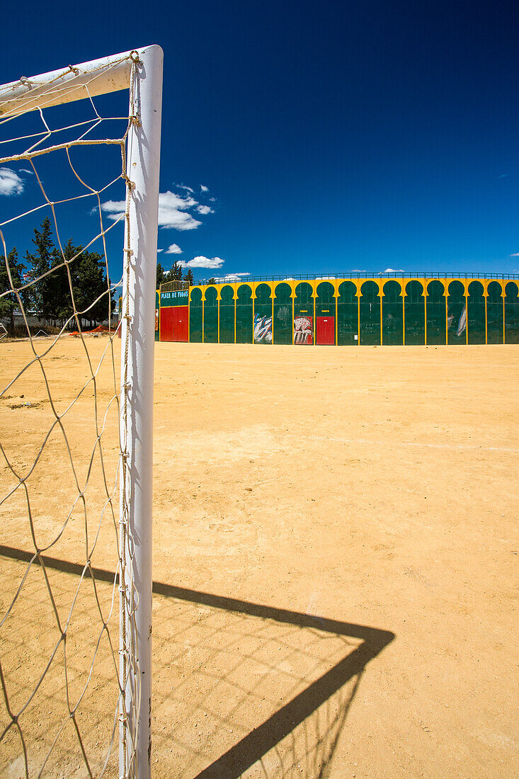 A unique view of a portable bullring set up in Aznalcazar, Seville, Spain, with a football goal in the foreground under a clear blue sky.