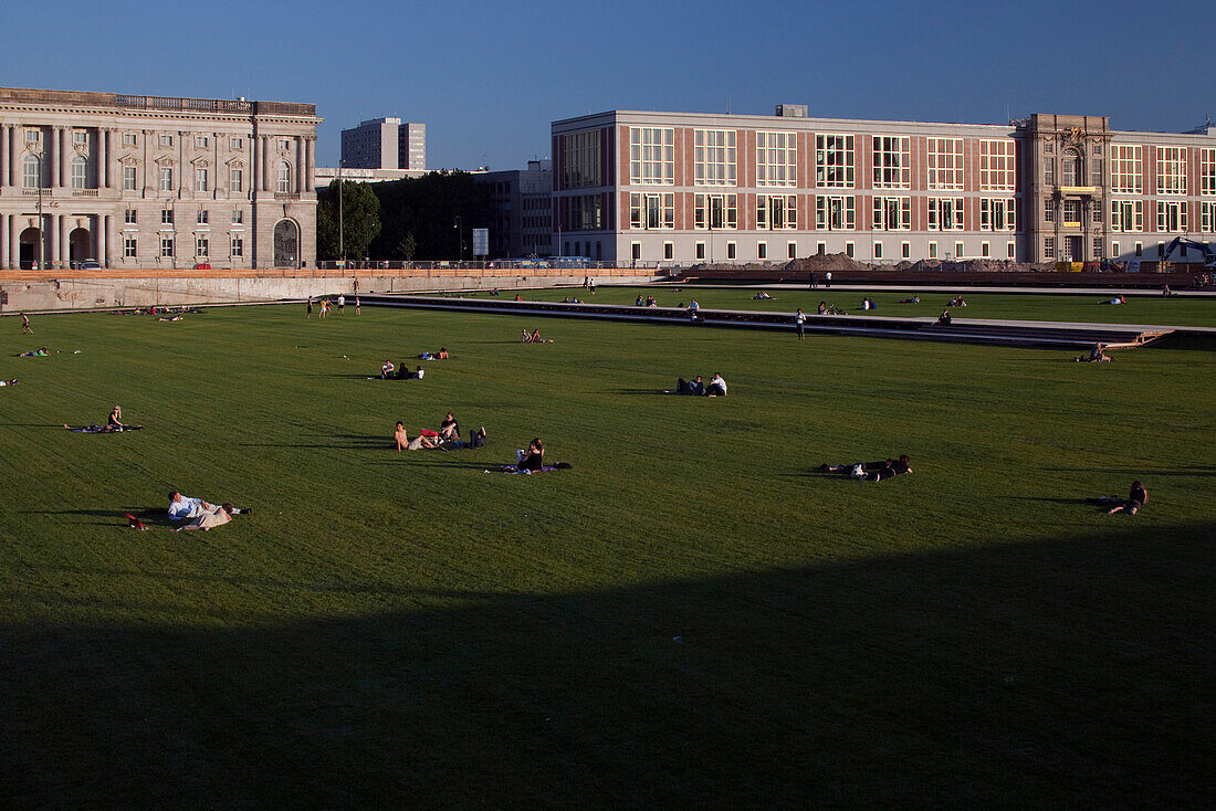 Visitors relax and sunbathe on the lush lawn of Schlossplatz, surrounded by historic buildings in Berlin on a bright day.