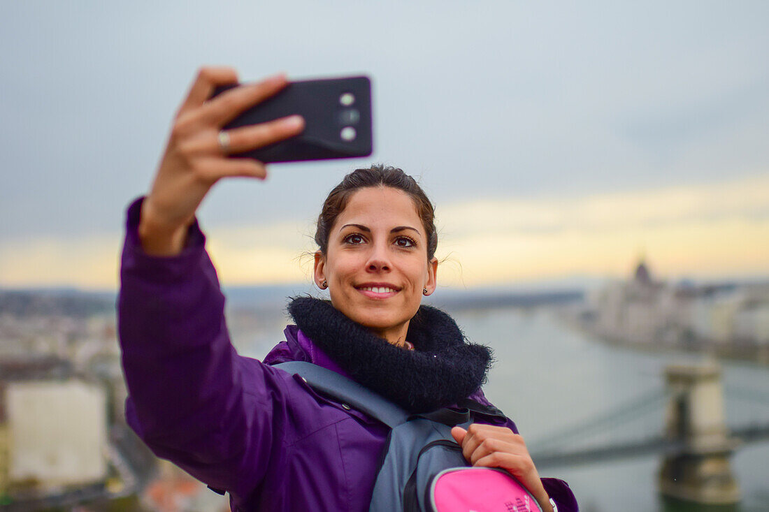 Young woman taking a selfie with skyline behind, including Parliament building and Danube River, Budapest, Hungary, Europe