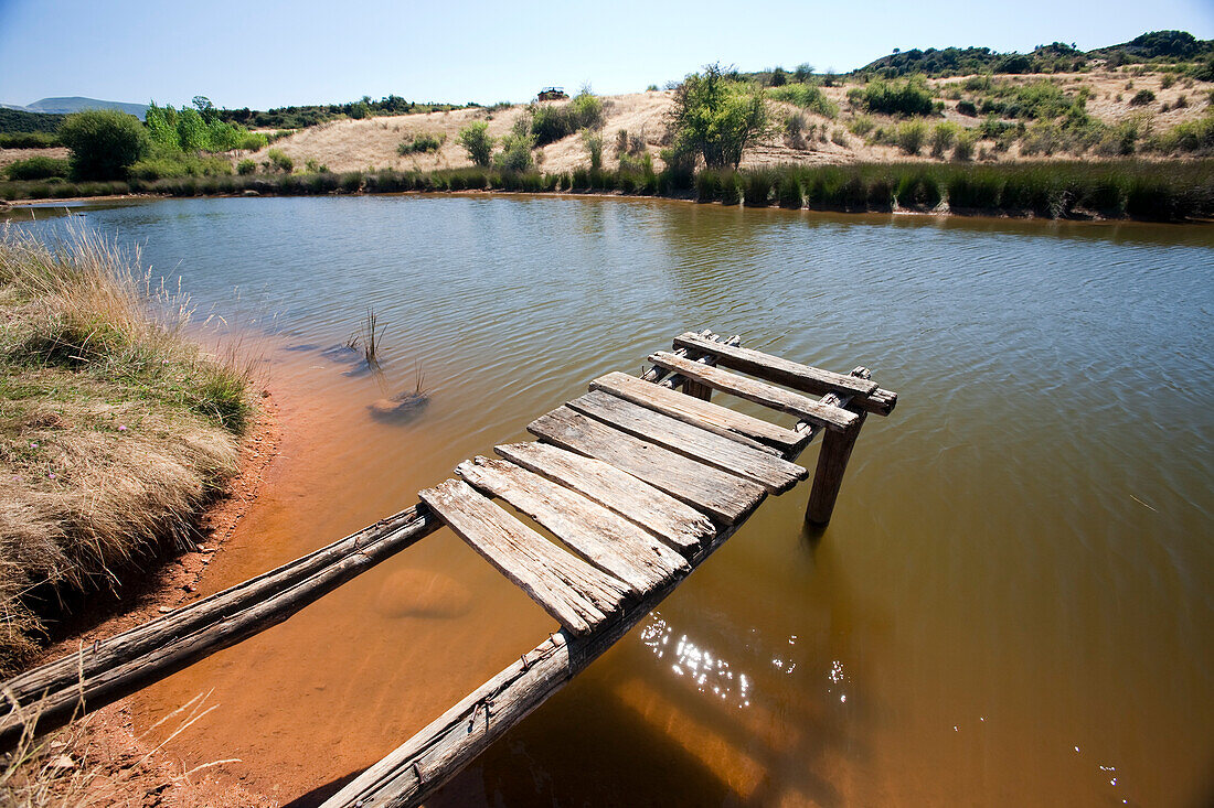 An old wooden dock extends into the tranquil waters of Lake Sumido, surrounded by the natural landscape of Las Médulas.