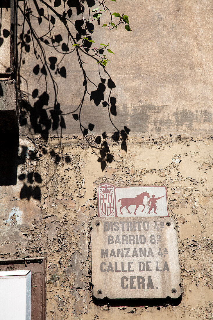 A weathered street sign identifies Placa de la Calle de la Cera in the vibrant Raval neighborhood of Barcelona under leafy branches.