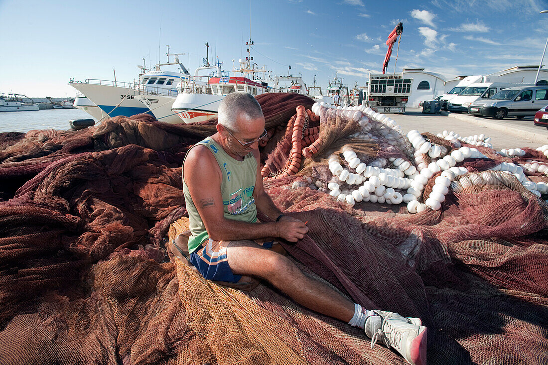 Seville, Spain, Aug 7 2008, A fisherman repairs his nets at Bonanza port in Sanlucar de Barrameda, Cadiz, while boats and vehicles surround him under a clear sky.